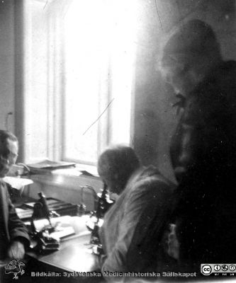 1928 års histologikurs i Lund
Fotoalbum märkt Histologikursen anno 1928. 
Detta album innehåller bilder från dåvarande histologiska avdelningen på anatomiska institutionen i Lund på Biskopsgatan 7. Liksom de övriga i serien skapades det av kursdeltagarna och tillägnades kursledaren professor Torsten Hellman som uppskattning av hans lärargärning. Bilden är inte namnsatt. Den visar professor Torsten Hellman i ljus laboratorierock vid ett kursmikroskop tillsammans med medicine studeranden i avdelningens mikroskoperingssal. Notera gärna att den elektriska lampan på mikroskoperingsbordet inte är tänd. Mikroskopen kunde använda fönstrets ljus.
Bildkälla Stiftelsen Locus Medicus Lundensis 250317.
Nyckelord: Locus Medicus Lundensis;Medicinska fakulteten i Lund;Histologiska avdelningen;Sydsvenska Medicinhistoriska Sällskapet;SMHS;Anatomiska Institutionen;Studenter;Lärare