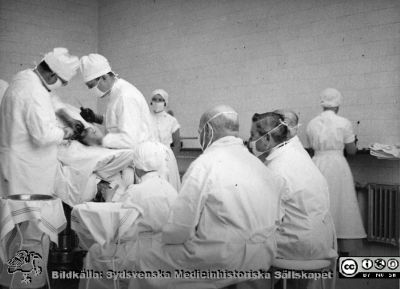 Operation på 1930-talet, troligen i Lund
Omonterat fotografi. Påskrift: "Från Elof Munck af Rosensköld under studietiden på 1930-talet. Aseptik."
Notera att besökare / åskådare visserligen har vita överdragskläder och munskydd, men det täcker inte nödvändigtvis näsan, och de har ingen mössa. Assisterande icke steril personal har däremot mössa. - Patienten ligger i s.k. Trendelenburgläge, dvs med huvudet lågt och benen högt. Narkossköterskan håller en öppen mask framför patientens ansikte; rimligen rör det sig alltså om narkos med eter eller möjligen kloroform.
Nyckelord: Kirurgi;Operation;Demonstration;Aseptik;1930-talet;Elof;Munck af Rosensköld;Foto;Omonterat;Kapsel 08