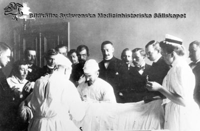 Professorn och kirurgöverläkaren Jacques Borelius opererar i gasljus i Lund 
Foto c:a 1900. Den första operationssköterskan, Sophiasystern Hedvig Olander assisterar. Kandidaterna bär mörk kostym. Bildkälla: Medicinhistoriska Museet i Lund.
Nyckelord: Jacques;Borelius;Professor;Gasljus;Operation;Kirurg;Kirurgi;Hedvig;Olander;Sophiasyster;Kandidater