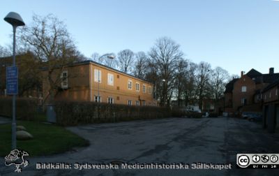 Universitetssjukhuset i Lund 2015 i solnedgång
Den gula baracken rymmer i bottenplanet lasarettets telefonväxel och på andra våningen diverse lokaler för olika entreprenörer verksamma på sjukhuset. Dess skönhetsvärde är näst intill obefintligt. Till höger Livets Museums ovanjordiska kulvert med mark- och parkservice (Green) i bottenplanet och restaurang Valvet längre bort.

Hade Region Skåne brytt sig om skönhetsvärden skulle har kunnat bli en lika fin park som den lite längre bort framför Universitetsbiblioteket som universitetet förvaltar. Nu visar sjukhuset bara upp en bakgård för turister och andra fotgängare som kommer från Lunds centrum.
Nyckelord: Lasarett;Lund;Universitet;Universitetssjukhus;USiL;Telefonväxel