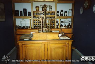 Apoteksreceptur på utställning
Medicinhistoriska Museet i Lund. Med hist samlingen 1992. I övrigt omärkt fotografi.
Enligt apotekare Greta Roslund, som ordnade utställningen, visar bilden apoteksutställningen på St Lars som den först ordnades av henne och apotekare Anna-Lisa Lagerlund. Efter hand kom det fram ytterligare material, bl.a. föremål från apoteket i Kävlinge. Utställningen gjordes till museets invigning 1992.
En skyddande glasvägg kom upp senare, men men före 1999. 
Det var egentligen 2 recepturer, som sammanställts, en vanlig med 2 arbetsplatser bredvid varandra och en kortare med 2 arbetsplatser mitt emot varandra. Man kan se 4 balansvågar, en på varje arbetsplats. 
Nyckelord: Apotek;officin;interiör;utställning;receptur;Våg;Balansvåg;Foto;Omonterat;Roslund;Greta;Anna-Lisa;Lagerlund;1990-talet;Kapsel 07