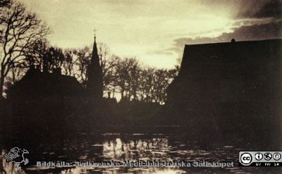 Helgonabacken i Lund med gård och märgelgrav. 
Allhelgonakyrkan i bakgrunden. Den stod färdig 1891. Foto rimligen från 1890-talet, i samma serie som andra bilder med samma motiv, av Lina Jonn. Dammen kan vara den som fortfarande finns utanför UB i Lund.
Nyckelord: Lasarett;Lund;Universitet;Universitetssjukhus;Kyrka;GÃ¥rd;Allhelgona;Helgonabacken