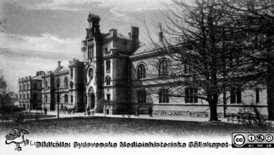 Fotografiskt vykort med 1867/68 års kirurgiska klinik i Lund från nordost. 
Notera att det står en hästskjuts utanför huvudentrén. Ögonkliniken (längst till vänster) stod färdig 1884. Många av träden är små, och bilden är därför troligen från 1800-talets slut. Från repronegativ i lasarettsfotografernas pärm "Gammalt A". Lund.
Nyckelord: Lund;Lasarett;Kirurgi;Klinik;Exteriör;Vykort;Universitet;Universitetssjukhus