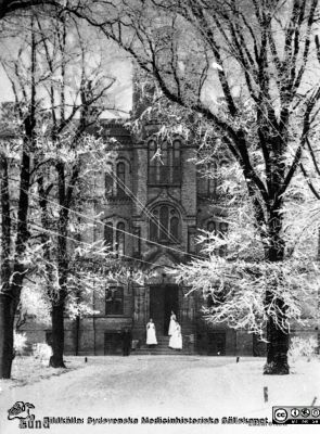 Vykort med entrépartient till 1867/68 års kirurgiska klinik i vinterskrud. 
Fotografen Lina Jonns Eftr. (Per Bagge) tog ett antal bilder av snarlik typ i början på 1900-talet. Från repronegativ i lasarettsfotografernas pärm "Gammalt A". Lund.
Nyckelord: Lund;Lasarett;Kirurgi;Klinik;Exteriör;Vykort;Universitet;Universitetssjukhus