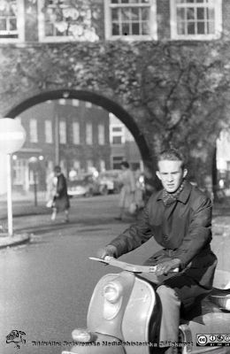 Höstlig vy 1961med en ung man på en scooter. 
Lasarettsfotograferna. Lund. Pärm "Div. tagningar, 1960 och t.v.". Lasarettets exteriörer, 1960-talet. Foto norrut mot valvet mellan medicinska (till vänster) och reumatologiska (till höger) klinikerna. Foto 1961. Från negativ
Nyckelord: Lasarett;Lund;Universitet;Universitetssjukhus;USiL;Medicin;Reumatologi;Valv;Scooter;Höst;Klinik