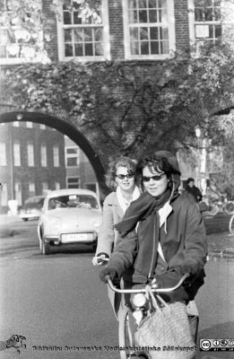 Höstlig vy 1961 med cyklister och en bil, norrut mot valvet mellan medicinska (till vänster) och reumatologiska (till höger) klinikerna. 
Lasarettsfotograferna. Lund. Pärm "Div. tagningar, 1960 och t.v.". Lasarettets exteriörer, 1960-talet. Vildvinet på valvet är avlövat och det ligger löv på marken närmast kameran. Foto 1961. Från negativ
Nyckelord: Lasarett;Lund;Universitet;Universitetssjukhus;USiL;Cyklist;Medicin;Reumatologi;Klinik;Valv;Höst