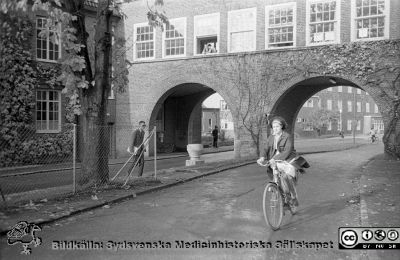 Höstlig vy 1961, norrut mot valvet mellan medicinska (till vänster) och reumatologiska (till höger) klinikerna. 
Lasarettsfotograferna. Lund. Pärm "Div. tagningar, 1960 och t.v.". Lasarettets exteriörer, 1960-talet. Vildvinet på valvet är avlövat och det ligger löv på marken närmast kameran. Foto 1961. Från negativ
Nyckelord: Lasarett;Lund;Universitet;Universitetssjukhus;USiL;Medicin;Reumatologi;Valv;Klinik;Höst;Cyklist