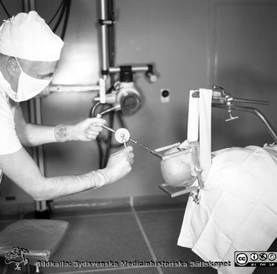 Lasarettet i Lund 1958. Professor Lars Leksell sätter en stereotaktisk ram på en patient
Lasarettsfotograferna. Lund. Pärm "Div. tagningar, 1960 och t.v.". Foto 1958. Professor Lars Leksell sätter en stereotaktisk ram på skallen på en patient. Här borrar han ett litet hål i skallbenets yttre skikt för ramens främre stift. Han skall sedan göra en encefalografi på patienten inför en stereotaktisk operation. Från negativ.
Nyckelord: Lasarett;Lund;Undervisning;Universitet;USiL;Neurokirurgi;Stereotaxi