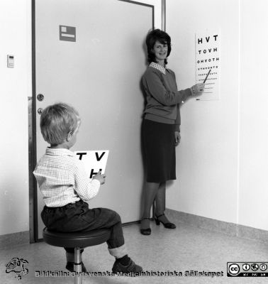 Ortoptisten Janet Hall Lindberg provar synskärpan på ett barn 
Sjukhusfotograferna i Lund. Pärm Svart-vita neg. 1987. 130/-87. Provningen gör med en s.k. HVOT-tavla i jämförelsevis enkel miljö och med undersökaren i egna kläder; som kanske på en förskola. Från negativ.
Nyckelord: Laboratorium;Lund;Universitetssjukhus;USiL;Ögon;Klinik;Oftalmiatrisk;Oftalmologisk;Barn