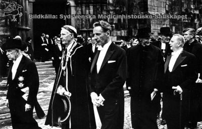 Akademisk festprocession 1968
Philip Sandblom (1903 - 2001), just lagerkransad professor i kirurgi vid Lunds Universitet från 1950, Rector Magnificus där 1957 - 1968. Här tillsammans med på sin högra sida f. universitetskanslern, f. landshövdingen mm Arthur Thomson och på sin västra landshövding Gösta Netzén vid uttåget ur domkyrkan i Lund efter en extra promovering i samband med universitetets firande av sitt 300-årsjubileum våren 1968. Philip Sandblom bär lagerkrans eftersom han just promoverats till filosofie hedersdoktor. Längre till höger i bild ses prorektor Per Stjernquist (i hög  hatt) och i samspråk med landstingsrådet Torsten Andrée, med halskedja. I nästa led ses universitetsrådet Stig Hammar till höger i bild, barhuvad.
Nyckelord: Kapsel 01;Regionarkivet;Lund;Fest;Procession;Promotion;Domkyrkan;Jubileum;