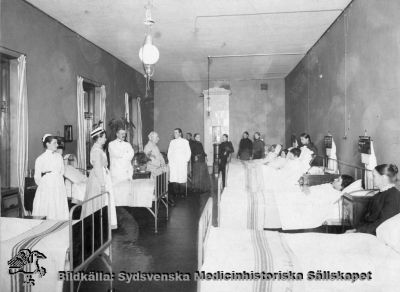 Läkarrond på kvinnlig sjuksal på "invärtes" (=medicinska) avdelningen på gamla lasarettet i Lund
Påskrift: "Den yngre av de båda läkarna är Karl Petrén. MS 7.297". Ingen ytterligare märkning.
Troligast Seved Ribbing i den lite tonade läkarrocken. Han bar ofta en sådan. Gasljuslamporna i taket talar för att kliniken ännu inte är elektrifierad, dvs rimligen är bilden tagen nära sekelskiftet 1900. Foto monterat på kartong. 
Nyckelord: Kapsel 02;Regionarkivet;Lasarettet;Lund;Medicin;Klinik;Gasljus;Seved;Ribbing;Rond;Läkarrond