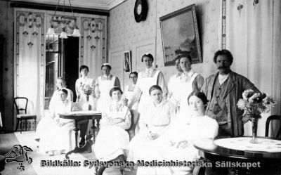 Dagrum klass 1 på Lunds Hospital
Fotografiskt vykort, aldrig avsänt. Påskrift: "Salongen 1 klass 1921". St Lars Hospital hade då en första klass-avdelning för bemedlade patienter som själva (eller deras släktingar) betalade för vården. Personerna på bilden är rimligen mest mentalvårdare och annan personal. Mannen finns på ett annat kort omnämnd som "Olsson som skötte Tranan och fåglarna i dammen". Det är oklart om han är vaktmästare eller patient.
Nyckelord: Kapsel 20;St Lars;Lund;Skötare;Mentalvårdare;Dagrum