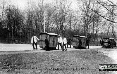 Matvagnar på den smalspåriga järnvägen på St Lars i Lund
Kapsel 22 med bilder från St Lars i Lund. Matvagnar. En vagn till varje paviljong. Skötare Ingvar Persson till höger. Sysselsättning. Fotografiskt vykort, rimligen från 1900-talets första decennier. Originalfoto. Publicerat på sid 99 i Truedsson 1929. Ej monterat. Spårvägen anlades 1890 i samband med att asylen (=västra sjukhuset) byggdes. Den lär ha lagts ned i slutet på 1930-talet. 
Nyckelord: Kapsel 22;St Lars;Lund;Personal;Skötare;Patienter;Mattransport;Järnväg