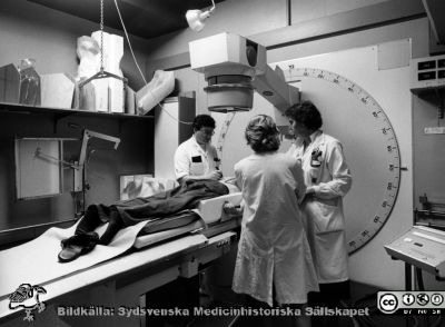 Onkologiska kliniken i Lund i slutet på 1980-talet. Högvoltsavdelningen. Simulator 
Foto Ola Terje låda A bild 17/2. Simulator för exakt inställning av strålfält. som ritas på patientens rygg. Sjukhusfysiker Inger Erlandsson längst till höger. Kanske Maria Albertsson med ryggen mot kameran.
Nyckelord: Terje;1980-talet;Onkologi;Interiör;Högvoltsavdelning;Strålkanon;Strålfält