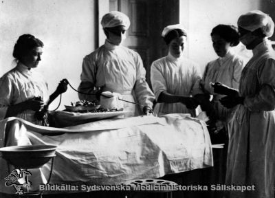 Bilden har använts som illustration vid utbildning i sterilitet
Påskrift: "Utbildning, sjuksköterskor." 
Rimligen foto från 1900-talets första halva, innan man hade börjat begära mössa och munskydd på alla i en operationssal.
Nyckelord: Sjuksköterskor;utbildning;1900-talet;omonterat foto;kirurgi;operation;operationssal