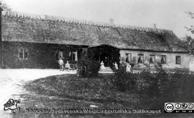 Kyrkoherden Carl Daniel Edvard Petréns prästgård i Skånes Halmstad
Kapsel 30. Påskrift: "Boningslängan I Halmstad prestgård, en sommardag, ett av 1890-talets allra första år. På verandan: Pappa, Gustaf; trappan: Lotten, mamma, Louise. Venster om verandan: Viktor, Jack. Höger: Edvard, Bror, Tina, Ebba". Prästgården beboddes av familjen Petrén. Reprofoto. Ej monterat.
Nyckelord: Kapsel 30;Präst;Familj;Gård