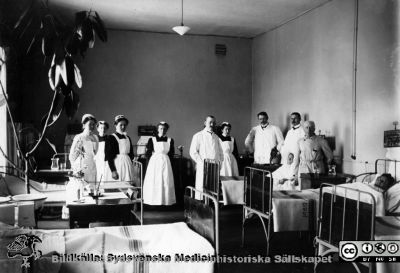  Professor Seved Ribbing går rond 1910 på medicinkliniken i Lund
Kapsel 30.  Originalfoto, monterat.Publicerad på sid. 199 i Flaum 1968. Bildtext där: "Nu finns elektrisk belysning och värmeledning. Kaminer och kakelugnar togs bort här år 1904. Avdelningssköterskan Laura Pedersen utexaminades med den första SSSH-kursen år 1905. Märk de svarta sängtavlorna, påskrivna med krita". 
Nyckelord: Kapsel 30;Överläkare;Professor;Lund;Medicin