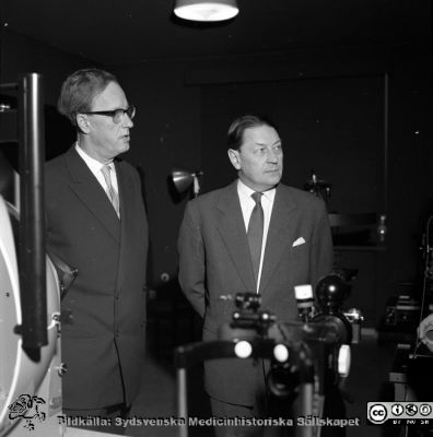 Prof Jan Waldenström och prof Karl-Erik Hogeman på ögonkliniken i Malmö. 
Album Äldre neg. 1951-1958 MAS i lasarettsfotograf Björn Henrikssons samling. Kanske i samband med invigningn av bl.a. ögonklinikens nya hus 1961-01-21. Apparaterna är en Goldmann-perimter och en Zeiss ögonbottenkamera.
Nyckelord: MAS;Malm;1951-1958;Direktion;Inspektion;gonklinik