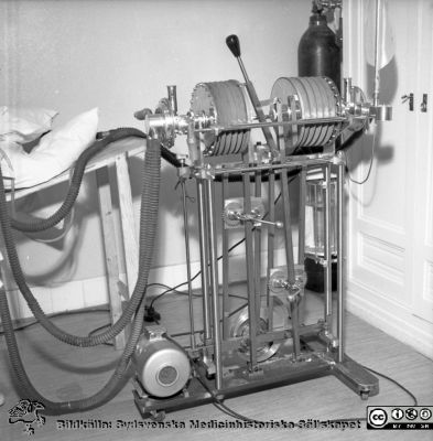 Prototyp till en Lundarespirator i klinisk prövning på infektionskliniken i Malmö den 19/5 1953. Från fotograf Björn Henrikssons samling
Negativpärm äldre 1951-58. MAS Malmö Allmänna Sjukhus. Epidemi. Respirator, en s.k. Lundiarespirator. 19/5, 53. Negativ.
Stig Cronberg 2012: Den allra första Lundiarespiratorn var en trampmaskin, ungefär som gammaldags symaskiner, men denna har fått en motor som driver den. Den har två bälgar så att man kan koppla två patienter till samma respirator. När den ena pumpas in, fylls bälgen för den andra patienten och vice versa. Till en början hade man olika konstruktioner och prövade sig fram. Just denna respiratortyp för 2 patienter vållade hygieniska problem och kom aldrig till serietillverkning. Senare fick Lundiarespiratorn ett metallhölje. I den som är på bilden ligger de mekaniska delarna helt öppna, vilket är både en fördel och en nackdel. Med sin enkla konstruktion var den mycket driftsäker och kunde användas var som helst.  I Köpenhamn använde man Lundiarespiratorn in på 1980-talet. Egentligen vore den idealisk för afrikanska förhållanden. Om någonting går sönder är det inte någon elektronikdetalj, som för dyra pengar måste införskaffas långt ifrån utan man kan vända sig till närmaste cykelmakare.
Nyckelord: Pärm äldre 1951-58;MAS;Malmö_;Respirator;Iinfektionsklinik;Epidemisjukhus