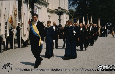 Processionen från universitetet vid doktorspromotion i Lund 1989
Bakom marskalken i blågult band (arkivarien Göran Larsson) och de två prestaverande pedellerna i blå långrockar skymtar Rector Magnificus Håkan Westling (halvskymd) och biskop Per-Olof Ahrén (med kors i halskedja) m fl. 
Nyckelord: Doktor;Doktorspromotion;Procession;Universitet;Promotion;Lund;Rector;Magnificus;Biskop