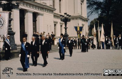 Processionen från universitetet vid doktorspromotion i Lund, troligast 1989
Medicinska fakultetens marskalk i grönt axelband i bakgrunden. Bakom henne kanske jubeldoktorn Sophus von Rosen. I förgrunden juridiska fakulteten med marskalk i blått band följd av juristernas hedersdoktorer Torstein Eckhoff och Tore Landahl.
Doktorn efter avlagda prov bör vara Sten Erik Holmberg.

Nyckelord: Doktor;Doktorspromotion;Procession;Universitet;Promotion;Lund