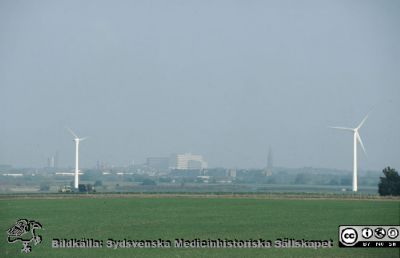 Vårligt soldis över lundaslätten vid Lunds Energis (nu Kraftringens) vindkraftverk vid Värpinge. 
Lasarettet i Lund i bakgrunden. Helikopterplattan på taket är ännu inte byggd. Kylvattentornet vid Lunds Energis (nu Kraftringens) kraftvärmeverk har byggts. Foto troligast på 1990-talet.
Nyckelord: Lasarett; Lund; Universitet; Universitetssjukhus; Exteriör;Lundaslätten