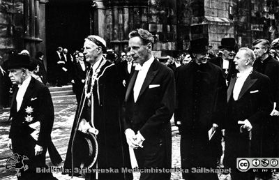 Processionen efter jubileumspromotionen vid universitetets firande av sitt 300-årsjubileum våren 1968.
Philip Sandblom (1903 - 2001), just lagerkransad professor i kirurgi vid Lunds Universitet från 1950, Rector Magnificus där 1957 - 1968. Här tillsammans med på sin högra sida f. universitetskanslern, f. landshövdingen mm Arthur Thomson och på sin västra landshövding Gösta Netzén vid uttåget ur domkyrkan i Lund efter en extra promovering i samband med universitetets firande av sitt 300-årsjubileum våren 1968. Philip Sandblom bär lagerkrans eftersom han just promoverats till filosofie hedersdoktor. Längre till höger i bild ses prorektor Per Stjernquist (i hög  hatt) och i samspråk med landstingsrådet Torsten Andrée, med halskedja. I nästa led ses universitetsrådet Stig Hammar till höger i bild, barhuvad.
Nyckelord: Kapsel 01 Regionarkivet;Lund;Universitet;Jubileum;Promotion