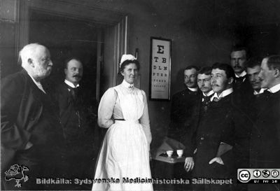 Ögonkliniken i Lund 1903. Professor Mikaël Löwegren, docent Albin Dahlén, översköterskan K. Theresia Löwenadler och tjänstgörande kandidater 
Foto på ögonkliniken i Lund. Se också bildfil 101014-002p. Påskrift på baksidan av denna bild: "De 3 främsta kandidaterna: Hirschlaff, Helsingborg. Westergård, f.d. Halmstad, Pallin Kristianstad. Den långe bakom: Wallengren, Landskrona. Översköterskan Fröken Löwenådler". Översköterskans efternamn är svårtytt. I databasen "Sveriges Befolkning 1900" från finns en översköterska Klara Theresia Löwenadler, född 1867 i Eksjö och skriven i Krafts rote i Lunds stadsförsamling. Påskrift på ramens baksida: "Nr 4 fr.v. är Herold Hirschlaff intygar sonen Erik Hjortsjö 16/11 -55". 
Fotot har också funnits i Sydsvenska Medicinhistoriska Sällskapets arkiv digitaliserades där c:a 2002 för Håkan Westling. Se fil 13Löwegren. Bilden anges där vara tagen 1903, och de tjänstgörande kandidaterna anges heta Gustaf Pallin, Emil Westergård, Herold Hirschlaff, Sven Johansson och Herman Wallengren
En mycket snarlik bild, rimligen från samma fototillfälle, finns publicerad i  'Ur en lundamedicinares minnesbok. Anteckningar gjorda av Michael Kolmodin Löwegren. Med kommentar utgivna av Ynge Löwegren'. Sydsvenska Medicinhistoriska Sällskapets Årsskrift 1971, sid 58 - 72. Fil 030205c.tif.
Nyckelord: Kapsel 02 Regionarkivet