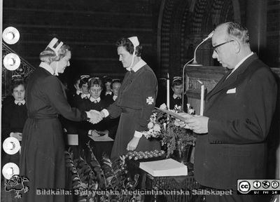 Kursavslutning vid Södra Sveriges Sjuksköterskehem (SSSH) ht 1956.
Iris Stålbåge (föreståndare på Södra Sveriges Sjuksköterskehem, SSSH) överlämnar brosch och diplom, assisterad av professor Gösta Glimstedt vid en högtidlig avslutning i Allhelgonakyrkan i Lund, höstkursen 1956. Den utexaminerade eleven är ej identifierad.
