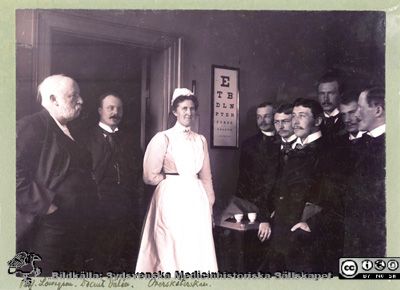 Professor Löwegren i ögonkliniken i Lund år 1903. 
Påskrift: "T.v. dåvarande docenten, sedermera professorn i oftalmiatrik Albin Dalén, t.h. tjänstgörande vid kliniken Gustaf Pallin, Emil Westergård, Herold Hisrschlaff, Herman Wallengren och Sven Johansson. Ur med. hist. sällsk. årsskrift 1971. Sid 67. MS 5.559. Lasarettsområdet söder interiör". - 
Sköterskan hette Karla Theresia Löwenadler, f. 1867 i Eksjö enligt påskrift på en annan kopia av denna bild, funnen 2010 på ögonkliniken i Lund (fil 101014-002.tif). Ett namn på de tjänstgörande saknas. Foto monterat på kartong.
Nyckelord: Kapsel 02;Regionarkivet;_ögonklinik;Oftalmiatrik;Oftalmologi;Översköterska