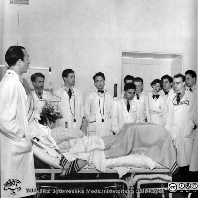 En undervisningdrond på medicinska kliniken i Lund 1950
Docent Nils Alwall på medicinska kliniken vid en patientsäng med en skara medicine kandidater. Från Bror Olsson, Erik Liljeroth och Bertil Wickman: "Ett bildverk om Lund". Allhems förlag, Malmö 1950. Fotograf: Erik Liljeroth. 
Nyckelord: Lasarettet;Lund;Universitetssjukhuset;USiL;Intern;Medicinska;Kliniken