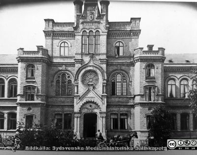 Dåvarande kirurgiska kliniken i Lund.
Fasad mot öster med huvudentrén. Hästskjutsen talar för att bilden är tagen under något av husets tidiga decennier. Det stod klart 1867. Växtligheten pekar på att huset är fotograferat åtminstone några år senare.  Bildkälla Denise Andersson 2021. Ur  hennes mans farfars efterlämnade bilder. Denne var Charly Urbanus Andersson, 18920525-3918, född i Ljunits, Sjörups församling vid Ystad, död 1974-02-14 och då kyrkobokförd på S Vallösa 5, 27012 Rydsgård. Han var skräddare och fotograf, bosatt i Vallösa utanför Ystad. 
Nyckelord: Kirurgisk klinik;Lasarettet i Lund;Universitetssjukhuset i Lund