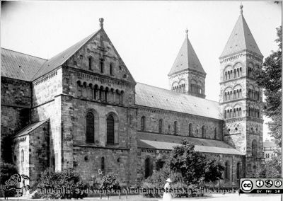 Domkyrkan i Lund, fasaden mot norr. 
 Bildkälla Denise Andersson 2021. Ur  hennes mans farfars efterlämnade bilder. Denne var Charly Urbanus Andersson, 18920525-3918, född i Ljunits, Sjörups församling vid Ystad, död 1974-02-14 och då kyrkobokförd på S Vallösa 5, 27012 Rydsgård. Han var skräddare och fotograf, bosatt i Vallösa utanför Ystad. Kvinnans och barnens kläder antyder att bilden troligen är tagen en högsommardag, kanske på 1930-talet.
Nyckelord: Lund;Domkyrka;Fasad mot norr.