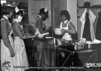 "Matvagnen göres i ordning till patienternas middag.".Tre unga kvinnor med elevmössor och ett vårdbiträde tan mössa men med enkel brosch på kragen. Bildkälla S. Anne-Marie Sörensen via S. Eva Ljunggren, SMHS.
Nyckelord: Utspisning;Lasarettet i Lund;Vårdavdelning