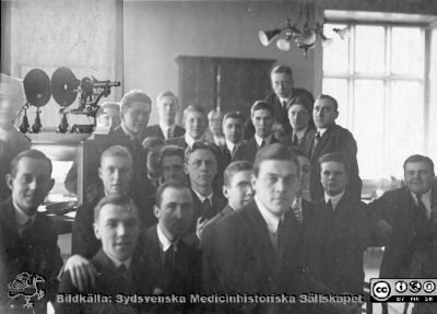 Thesaurus histologiae 1926
Fotoalbum på första bladet märkt 
"Thesaurus histologiae. 
thet är 
Een obiektiv beskrifning aff hendelser på then histologiska skåådebanan A.D. MDMXXVI. Med tacksamhet och vördnad överräckt av Kurs och amanuenser.". 
Detta album innehåller bilder från histologiska avdelningen 
på anatomiska institutionen i Lund på Biskopsgatan 7. Bildtext: "Fig. 19 "Före slaget." Rimligen kursfoto i mikroskoperingssalen på histologiska avdelningen på anatomiska institutionen. Notera gärna den ålderdomliga apparaten för projektion av mikroskopibilder i bildens övre vänstra del, rimligen ett hemmabygge på en klassisk optisk bänkskena.
Bildkälla Stiftelsen Locus Medicus Lundensis 250317. Fotograf okänd. Studenten i bildens vänstra del och under apparaten skulle kanske kunna vara Göte Österlind (1907 - 1981), disputerad i histologi 1941 och sedermera överläkare i oftalmiatrik vid Malmö Allmänna Sjukhus från 1951. Han blev 1969 den förste professorn där när den tjänsten inrättades.
Nyckelord: Histologiska avdelningen;Locus Medicus Lundensis;Locus Medicus Lundensis;Medicinska fakulteten;Medicine studerande;Anatomiska Institutionen;Mikroskop;Projektor