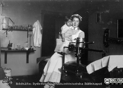 En ung sjuksköterska sitter på ett undersökningsbord med ett litet barn i famnen.
Bild 275 i husmor Anna Landelius (1880-1964) efterlämnade fotoalbum. Ingrid Frennhoff (f. 1944) hade övertagit detta album efter sin morfar som i sin tur tagit hand om det efter sin syster husmor Anna på Lasarettet i Lund. Gåva till Sydsvenska Medicinhistoriska Sällskapet nyåret 2020 - En ung sjuksköterska med SSSH-mössa och brosch sitter på ett undersökningsbord med ett litet barn i famnen. Kanske är det ett röntgenbord, för det sitter en dåtida typ av hållare för röntgenrör på det. Något röntgenrör syns dock inte i hållaren.
Nyckelord: SSSH;Södra Sveriges Sjuksköterskehem;Röntgen;Undersökningsbrits