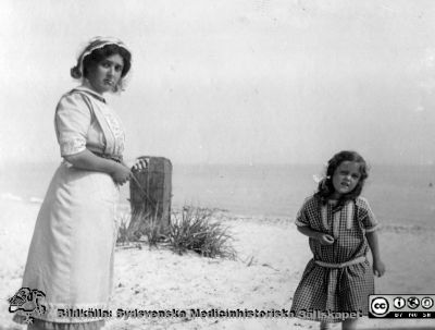 Kvinna med barn på sandstrand vid öppen kust. 
Bild 83 i husmor Anna Landelius (1880-1964) efterlämnade fotoalbum. Ingrid Frennhoff (f. 1944) hade övertagit detta album efter sin morfar som i sin tur tagit hand om det efter sin syster husmor Anna på Lasarettet i Lund. Detta album är en gåva från Ingrid Frennhoff till Sydsvenska Medicinhistoriska Sällskapet nyåret 2020.
Rimligen anhöriga till Anna Landelius. Kanske nära Falsterbo?
Nyckelord: Sandstrand;Barn