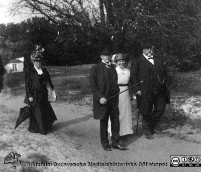 Två  män och två kvinnor på promenad. Anhöriga till Anna Landelius.
Bild 85 i husmor Anna Landelius (1880-1964) efterlämnade fotoalbum. Ingrid Frennhoff (f. 1944) hade övertagit detta album efter sin morfar som i sin tur tagit hand om det efter sin syster husmor Anna på Lasarettet i Lund. Detta album är en gåva från Ingrid Frennhoff till Sydsvenska Medicinhistoriska Sällskapet nyåret 2020.
Alla i bilden är väl klädda. Den bakre kvinnans väldiga hatt antyder att den är gjord nära sekelskiftet 1900. Den ljust klädda yngre kvinnans kläder är från ett senare decennium. Alla på bilden är troligast nära anhöriga till Anna Landelius. Kanske foto på hennes föräldrahem i Ljungsbro.
Nyckelord: Promenad