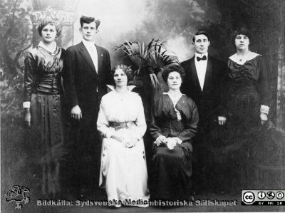 John och Augusta Persson i New York nära 1910-1914.
Enligt följebrev 17 feb 1989 från dir. Rune Rasmusson på svenska IBM, rimligen till professor Nils Oskar Berg, ordförande i styrelsen för John och Augusta Perssons stiftelse:
" Bäste Nils, Tack för senast. Det var ett mycket trevligt evenemang. Allt fungerade mycket bra. Bif. som lovat ett foto sedan omkring 1910 med John och Augusta Persson tillsammans med syskonbarn i New York. - Med bästa hälsningar Rune Rasmusson".
Det är Augusta som sitter till höger i mörk klänning och bredvid henne står John med mörk fluga i halsen. 
Etnologen Katarina Ek-Nilsson i Uppsala skrev 240927: "Längst till vänster står Emelia Andersdotter. Hon var syster till min mormor och emigrerade till NY 1912. Strax före 1920 återvände hon, blev bondhustru och ganska tidigt änka. Hon flyttade sedan hem till sin mamma (alltså en syster till Augusta). Jag har tydliga minnen av henne... 
Därefter en man, vars namn jag inte vet, men antagligen Kempf, då, eftersom nästa person är Gladys, kusin till mormor. Mormor pratade ofta om en Gladys. 
Sedan Augusta och John, och slutligen min mormor Anna Andersdotter... 
Året måste vara 1914 eller senare, för mormor var i NY 1909-1912. Därefter åkte hon hem (antagligen för att hjälpa till med småsyskon hemma), Emelia åkte iväg, och 1914 kom mormor åter till NY. Hon arbetade liksom Augusta i välbeställda familjer. ".
Nyckelord: Släktingar;Donatorer;Stiftelse;Lunds universitet;Medicinska fakulteten