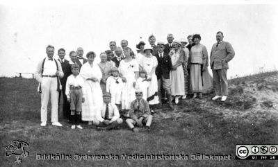 Erik M. P. Widmarks vänner på födelsedagsfest .
Enligt Eva Marie Widmark är personerna: längst t.h. Aron Westerlund; 5. från höger Gerda Johansson ; 8. från höger Erik Widmark ; 11. från höger Nora Widmark med pojkarna.
Påskrift på bildens baksida med delvis svårtydd handstil:
Löderups strandbad. Badgäster, födelsedagsfirare. 
Nora  i mitten m PH o CE. Erik längre bort t.h. 
Bredvid t.h. om Erik är Joh. Johansson m Gerda framför i vit hatt / sjal.
Aron Westerlundh längst t.h.?
Rimligen är det Noras födelsedag den 22 juli som firas.Initialerna PH och CE står för sönerna Per-Henrik (f. 1917) och Carl-Erik (f. 1920) som hon håller i händerna. Fotot är därmed rimligen taget i slutet på 1920-talet.
Foto privat. Fotograf okänd. Bildkälla Eva Marie Widmark.
Nyckelord: Badgäster;Fest;Födelsedagsfirare