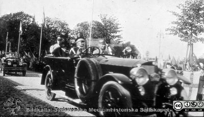 Kungligt besök på Ängelholms lasarett nära 1930. 
Efter diabild i sjuksköterskan Lillie Börjessons  samling från Ängelholms sjukhus. En stor öppen bil körs av en mycket guldgalonerad chaufför. Bilen har en kungakrona framför kylaren, mellan de stora lyktorna. Möjligen sittert Prins Bertil i det vänstra framsätet? Kanske är det kung Gustaf V som man ser i baksätet, vinkande. På andra sidan vägen och till höger står åskådare med flaggor, och en svensk flagga hänger på en stång till höger i bilden. Gråskalan i flaggorna är omvänd, dvs de blå fälten är ljusare än det gula korset. Bilden är alltså tagen med s.k. ortokromatisk film, som avbildade den svenska flaggan så. Den ersattes i början av 1930-talet av pankromatisk film som avbildar flaggan rätt. Bilden bör alltså vara tagen senast i början på 1930-talet. 
Bakom kommer en bil med registreringsskylt A4, dvs också den en kunglig bil. Notera att denna bil är vänsterstyrd, dvs troligen av brittiskt fabrikat.
Ängelholm fick ett epidemisjukhus på 1920-talet, och kanske härstammar bilden från dess invigning.
Nyckelord: Lasarett;Kungar;Kunglig;Bil;Automobil;Sjukhus;Engelholm;Ängelholm;Prins;Monark