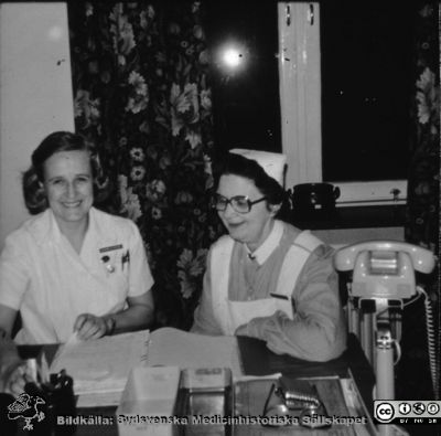 Ängelholms lasarett.  Två okända sköterskor vid ett skrivbord 
Efter en diabild i Lillie Börjessons medicinhistoriska bildsamling på Ängelholms lasarett.  Två okända sköterskor vid ett skrivbord på Lasarettet  Namnbroscherna är inte läsbara. Glasögonen talar för 1970- eller 1980-tal.
Nyckelord: Lasarett;Engelholm;Ängelholm;Sköterskor