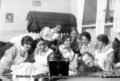 En grupp med ung vårdpersonal samlade runt en akustisk vevgrammofon. 
Troligen elever på SSSH (Södra Sveriges Sjuksköterskehem). Grammofontypen var vanlig på 1930- och 1940-talen innan de blev elektriska. Från Syster Elinas fotoalbum (Elina Holmberg?) .
