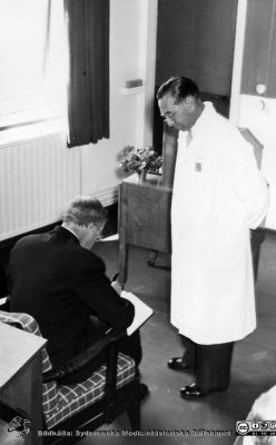 Kungligt besök på radiologiska kliniken i Lund 1951. 
Kung Gustaf VI Adolf skriver i radiologiska klinikens gästbok medan klinikchefen docent Bertil Ebenius står bredvid. Från Syster Elinas fotoalbum (Elina Holmberg?).
Nyckelord: Onkologisk;Radiologisk;Klinik;Lasarettet i Lund;Medicinska fakulteten;Invigning;Koboltkanon