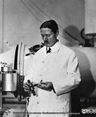 Professor Torsten Thunberg framför sin barospirator, en speciell andningsapparat han konstruerat och fått byggd.
Bildkälla: preparator Olle Hammar. 1920-talet	Professor Torsten Thunberg framför barospiratorn. Apparaten fick bara begränsad klinisk användning. Också publicerad i Westling: Tre professorer i fysiologi i Lund.
Nyckelord: Fakultet;Fysiologisk;Institution;Respirator;Medicinsk;Lund;Universitet