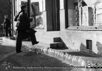  Svårtolkat studentskämt i Docentgatan 1958.
Mjölkflaskor blockerar en affär. Svårtolkat studentskämt i Docentgatan 1958. Kanske ditsatt av en ångerfull utfestad student? TetraPak hade visserligen redan 1952 börjat tillverka och sälja mjölk i pappförpackningar i Lund, men mjölken distribuerades fortfarande oftast i glasflaskor. Somliga var runda och andra var som man kan ana mer fyrkantigt moderna. Nu 2022 säljs all mjölk och många andra drycker i papp- eller plastförpackningar.
Nyckelord: Lunds studentkår.Mjölkflaskor.