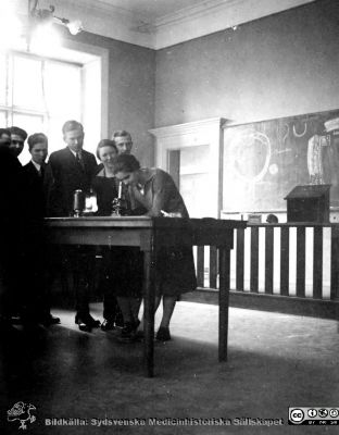 Thesaurus histologiae 1926
Fotoalbum på första bladet märkt 
"Thesaurus histologiae. Det är een obiektiv beskrifning aff hendelser på Den histologiska skåådebanan A.D. MDMXXVI. Med tacksamhet och vördnad överräckt av Kurs och amanuenser.". 
Detta album innehåller bilder från histologiska avdelningen 
på anatomiska institutionen i Lund på Biskopsgatan 7. Bildtext Fig 2. Kö". Studenterna skall titta på något i ett mikroskop.
Foto troligen i föreläsningssalen.
Bildkälla Stiftelsen Locus Medicus Lundensis 250317. Fotograf okänd.
Nyckelord: Anatomiska Institutionen;Histologiska avdelningen;Medicinska fakulteten i Lund;Universitetet;Medicine studerande;Amphioxer;Föreläsningsal