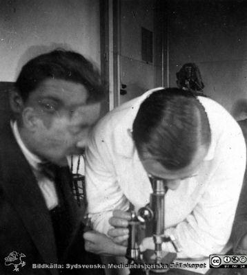 1928 års kurs i histologi i Lund
Fotoalbum märkt Histologikursen anno 1928. 
Detta album innehåller bilder från dåvarande histologiska avdelningen på anatomiska institutionen i Lund på Biskopsgatan 7. Liksom de övriga i serien skapades det av kursdeltagarna och tillägnades kursledaren, professor Torsten Hellman, som uppskattning av hans lärargärning. Bilden visar en av kursamanuenserna, "Eonen", Henry Dahlin enligt bildtext. Han hjälper studenten NN Linderoth vid dennes kursmikroskop.
Bildkälla Stiftelsen Locus Medicus Lundensis 250317.
Nyckelord: Locus Medicus Lundensis;Medicinska fakulteten i Lund;Histologiska avdelningen;Sydsvenska Medicinhistoriska Sällskapet;SMHS;Anatomiska Institutionen;Studenter;Lärare