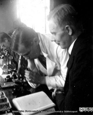 1928 års kurs i histologi i Lund
Fotoalbum märkt Histologikursen anno 1928. 
Detta album innehåller bilder från dåvarande histologiska avdelningen på anatomiska institutionen i Lund på Biskopsgatan 7. Liksom de övriga i serien skapades det av kursdeltagarna och tillägnades kursledaren, professor Torsten Hellman som uppskattning av hans lärargärning. Bilden visar en av kursamanunerserna, "Eonen", Henry Dahlin när han hjälper studenten Harry Johansson vid dennes kursmikroskop.
Bildkälla Stiftelsen Locus Medicus Lundensis 250317.
Nyckelord: Locus Medicus Lundensis;Medicinska fakulteten i Lund;Histologiska avdelningen;Sydsvenska Medicinhistoriska Sällskapet;SMHS;Anatomiska Institutionen;Studenter;Lärare