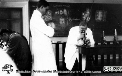 Foton från histologkursen i Lund 1929. 
Fotoalbum tillägnat Professor Torsten Hellman med tacksamhet och vördnad från histologkursen 1929. Bildtext "Chefen". Torsten Hellman vid mikroskopet framför svarta tavlan. Olof Ask  står avvaktande bredvid.
Nyckelord: Anatomiska Institutionen;Histologiska avdelningen;Föreläsningssal